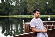 A young man in a stylish shirt leans thoughtfully on a wooden railing by a lake.