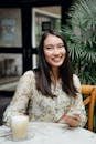 Cheerful young Asian lady with mobile phone in casual clothes smiling and drinking latte while chilling in outdoor cafe